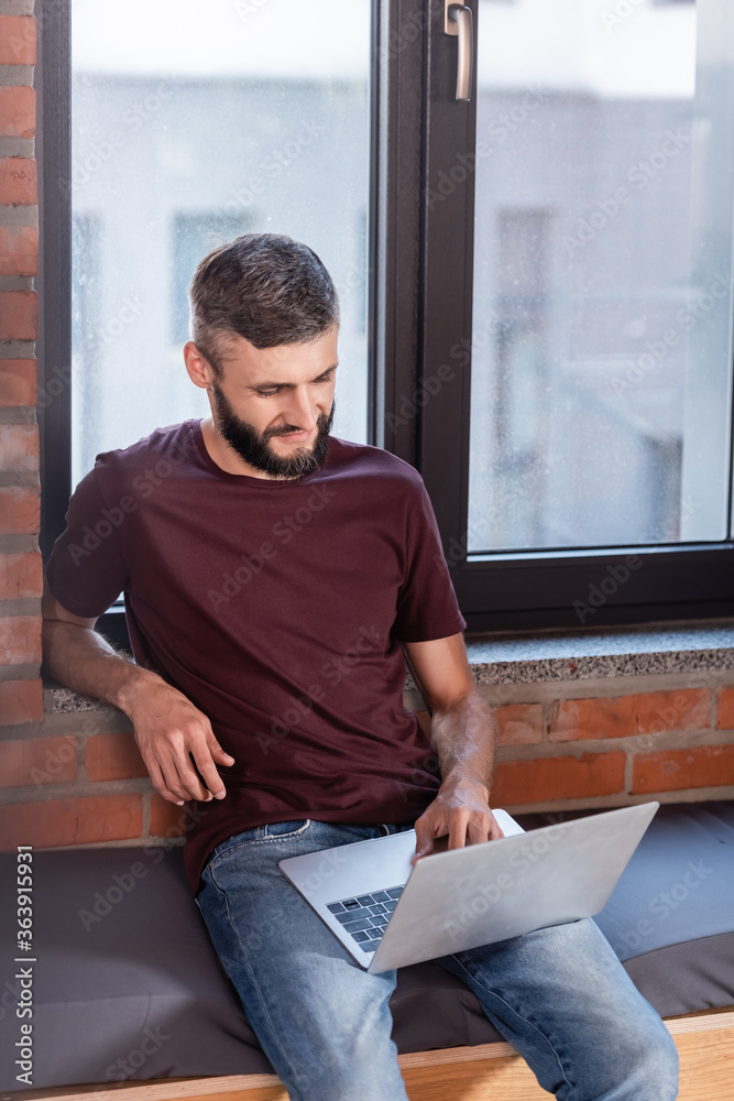 bearded businessman sitting on window bench and using modern laptop
