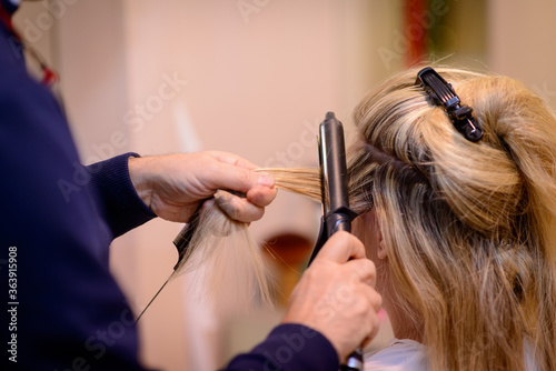 Mani di parrucchiera che con il ferro per capelli arriccia una ciocca di capelli biondi lunghi