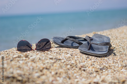 Selective focus on old slippers. Next are sunglasses. They are located on the yellow sand on the beach and it is a beautiful sunny day. Copy space.