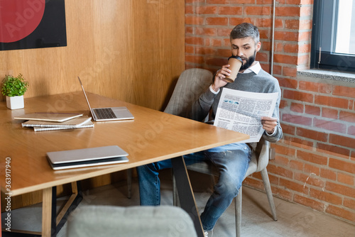 bearded businessman holding paper cup and reading newspaper near laptops © LIGHTFIELD STUDIOS