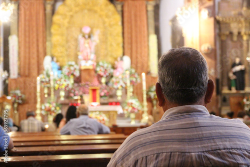 The back of an anonymous, elderly man, sitting in a nicely decorated Mexican church. A ceremony is being held. The catholic altar is very brightly endorsed with the statue of Jezus Christ.