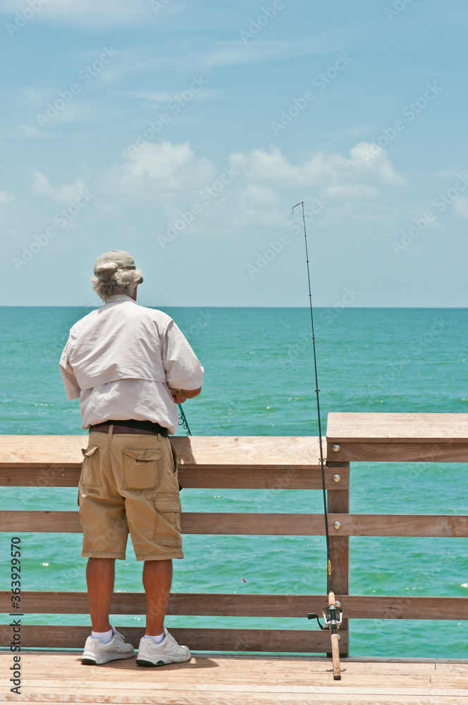 Back view, medium distance of a senior male fishing off a wood pier in the tropical waters of the gulf of Mexico on a sunny day Back view, medium distance of a senior male fishing off a wood pier in the tropical waters of the gulf of Mexico on a sunny day