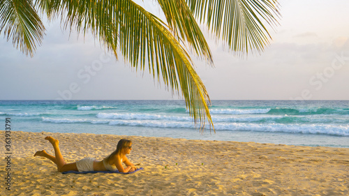 Young Caucasian woman lies on the beach and watches the surfers catching waves.