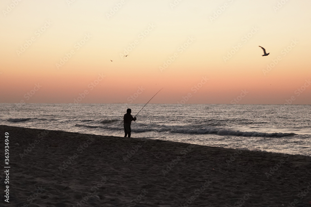 Fisherman catches fish from the shore at sunset. Old man holds fishing rod in his hands while standing on the sea coast. Dark male silhouette near water.