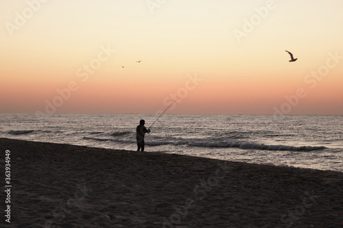 Fisherman catches fish from the shore at sunset. Old man holds fishing rod in his hands while standing on the sea coast. Dark male silhouette near water.