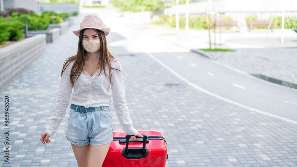 A beautiful young Caucasian woman in a medical mask walks down the street with a red suitcase. A girl with luggage travels alone during the height of the coronavirus epidemic. The empty city. A beautiful young Caucasian woman in a medical mask walks down the street with a red suitcase. A girl with luggage travels alone during the height of the coronavirus epidemic. The empty city.