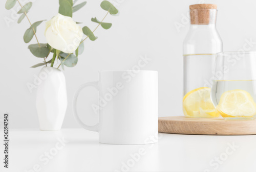 White mug mockup with flowers in a vase, glass and a bottle on a white table.