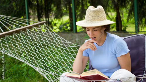 Pretty young woman reading interesting book in a hammock in green garden during weekend.Thoughtful curly brunette girl in summer straw hat absorbed in reading best seller sitting on rope hummock