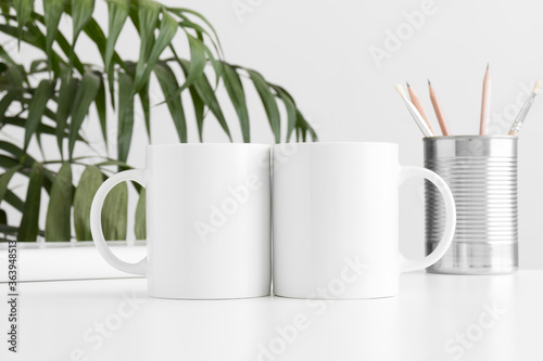 Two mugs mockup with workspace accessories on a white table and a palm plant.