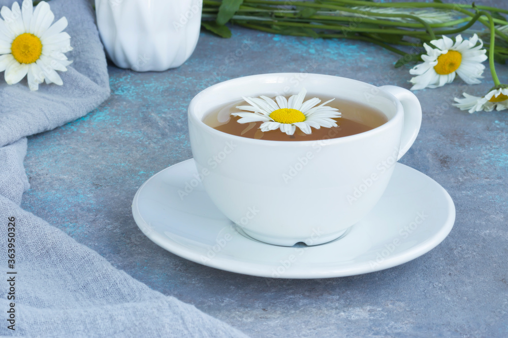 Herbal chamomile tea with fresh chamomile flowers in a white cup and saucer on a gray background. Bouquet of flowers on a wooden board and linen napkin. Herbal chamomile tea with fresh chamomile flowers in a white cup and saucer on a gray background. Bouquet of flowers on a wooden board and linen napkin.