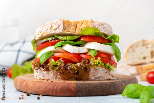 Big tasty sandwich of ciabatta with mozzarella cheese, tomatoes, basil and lettuce on the table. Sandwich with cheese and vegetables on a light background, vegetarian food. Italian Cuisine.