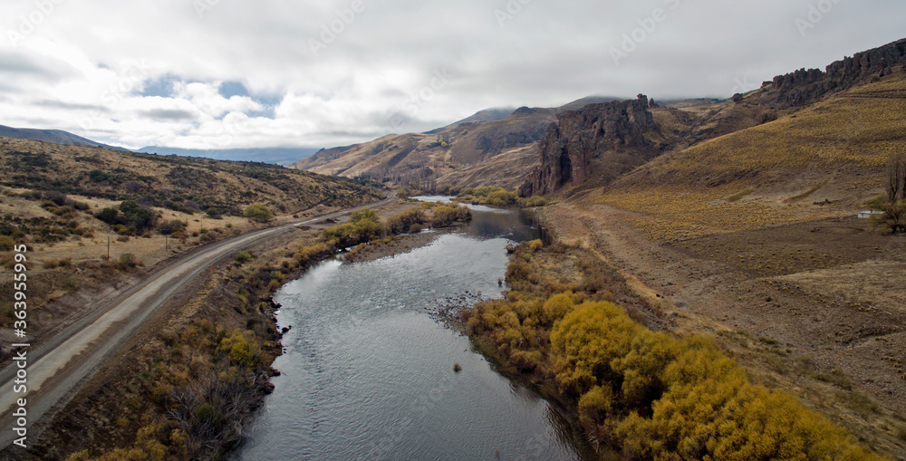 Enchanting valley. Aerial view of the rural dirt road across the golden meadow in autumn. The pure water stream flows across the prairie, mountains and rocky formations. 