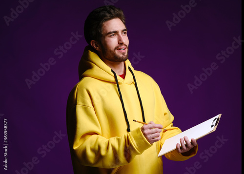 A young man of 25-30 years in glasses and a yellow sweatshirt holding a clipboard with clip in one hand and a pencil in the other