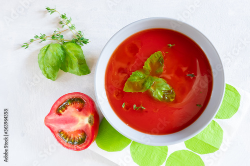 Top view of cold gazpacho soup in a grey bowl with half of a tomato and herbs on a light grey background