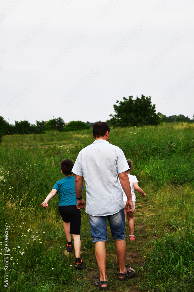 A friendly family, father holds the hand of a son and daughter, they walk together in a green meadow. A boy and a girl with his father go along the road and go into the distance. A friendly family, father holds the hand of a son and daughter, they walk together in a green meadow. A boy and a girl with his father go along the road and go into the distance.