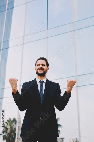 An excited man with winner spirit celebrating success, happy businessman with his arms raised standing near office building, overjoyed businessman raising his arms in victory outside a office building