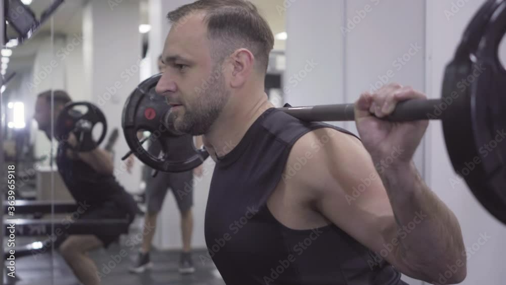 Live camera follows Caucasian sportsman lifting hand weights. Side view portrait of young handsome man exercising shoulder and back muscles in gym. Personal trainer controls body-builder training.