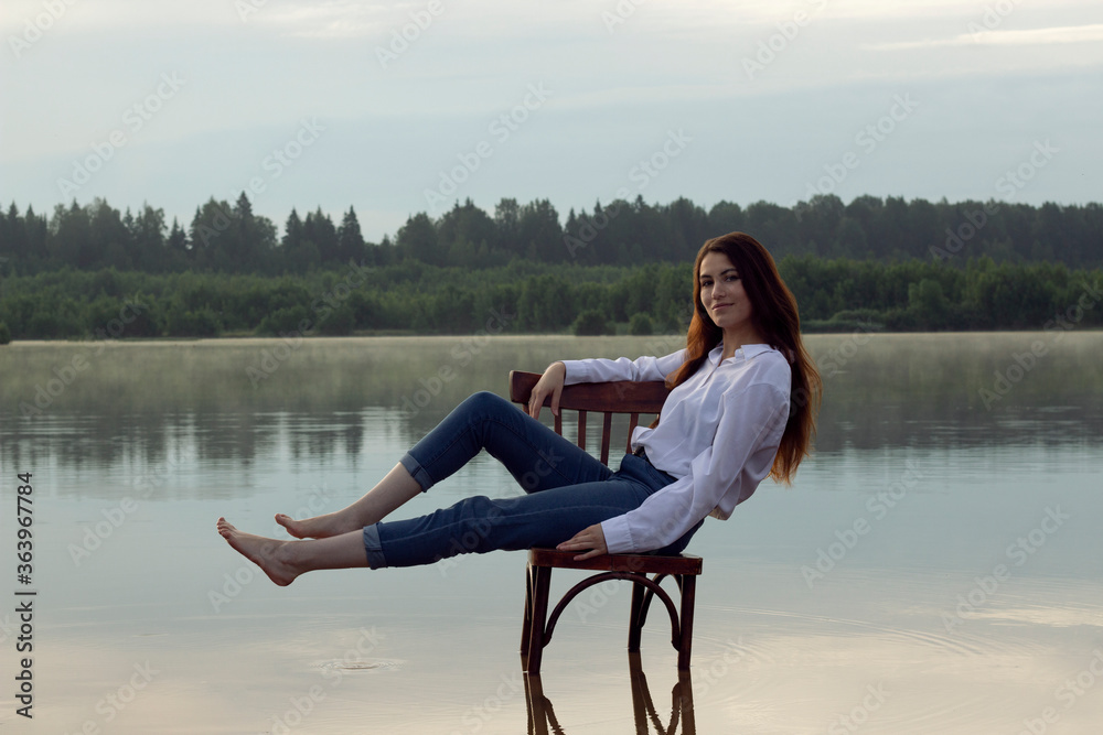 A girl in jeans and a white shirt, sitting on an old chair in the middle of the lake. A girl with long hair in the middle of the lake.