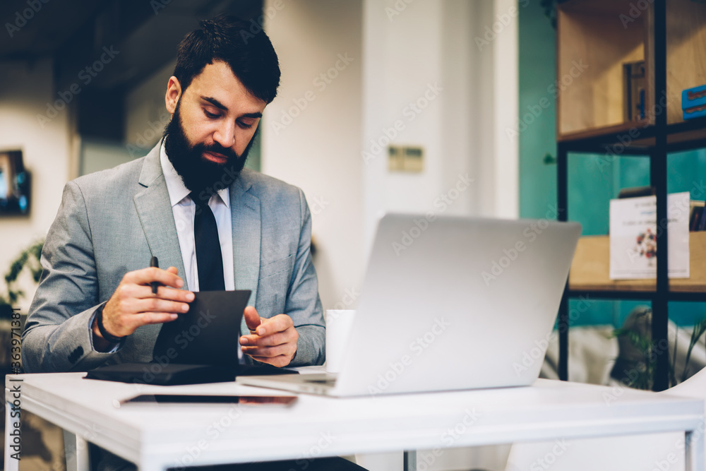 Bearded businessman dressed in formal wear checking documentation sitting at desktop with laptop computer connected to wireless internet in office interior near copy space area for advertsing Bearded businessman dressed in formal wear checking documentation sitting at desktop with laptop computer connected to wireless internet in office interior near copy space area for advertsing