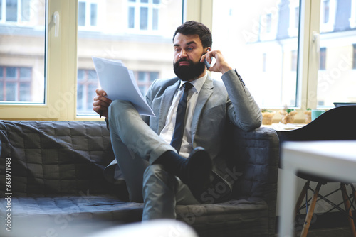 Concentrated bearded male owner of trading corporation discussing with secretary assistant accounting report of income while sitting in office, businessman proofreading contract calling to lawyer