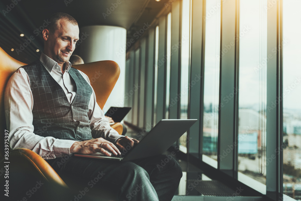 A cheerful successful businessman is working on his laptop while sitting on an armchair indoors of a luxurious office interior, with a window and a copy space zone on the left for an advertising text - obrazy, fototapety, plakaty 