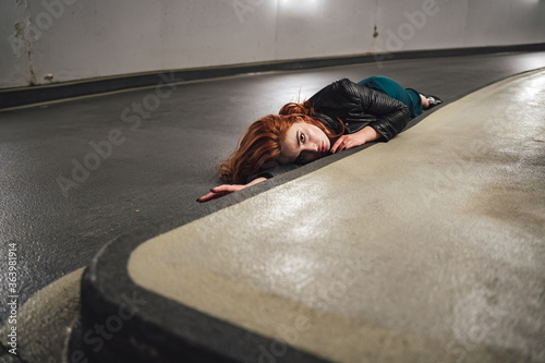 Red haired female model laying on floor inside parking house along curb. Young woman with open eyes acting dead victim of accident.