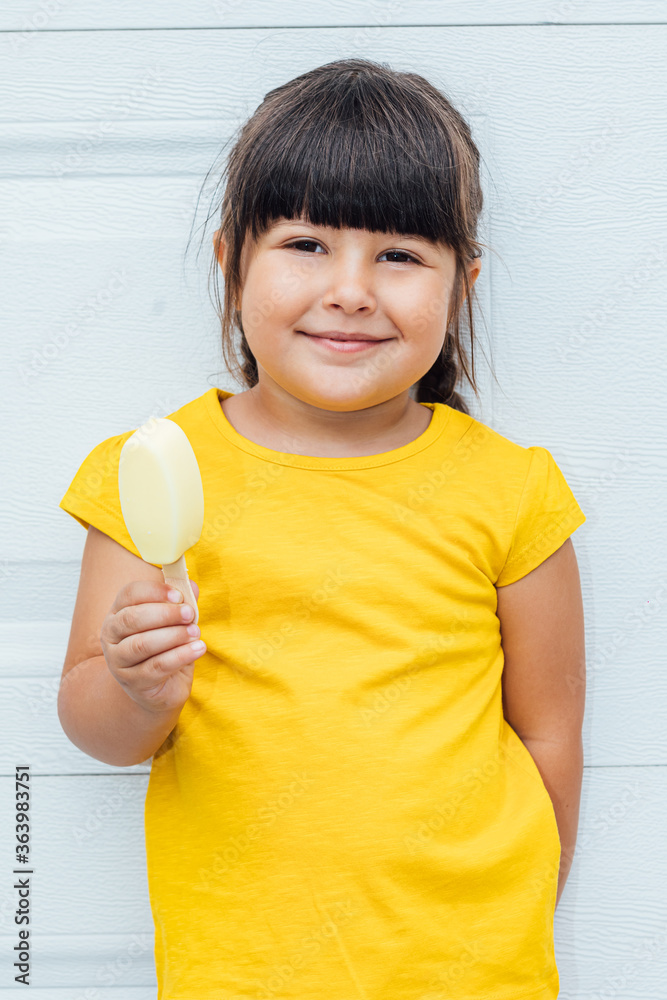 Adorable little girl with black hair eating ice cream, wearing a yellow shirt leaning against white background Adorable little girl with black hair eating ice cream, wearing a yellow shirt leaning against white background