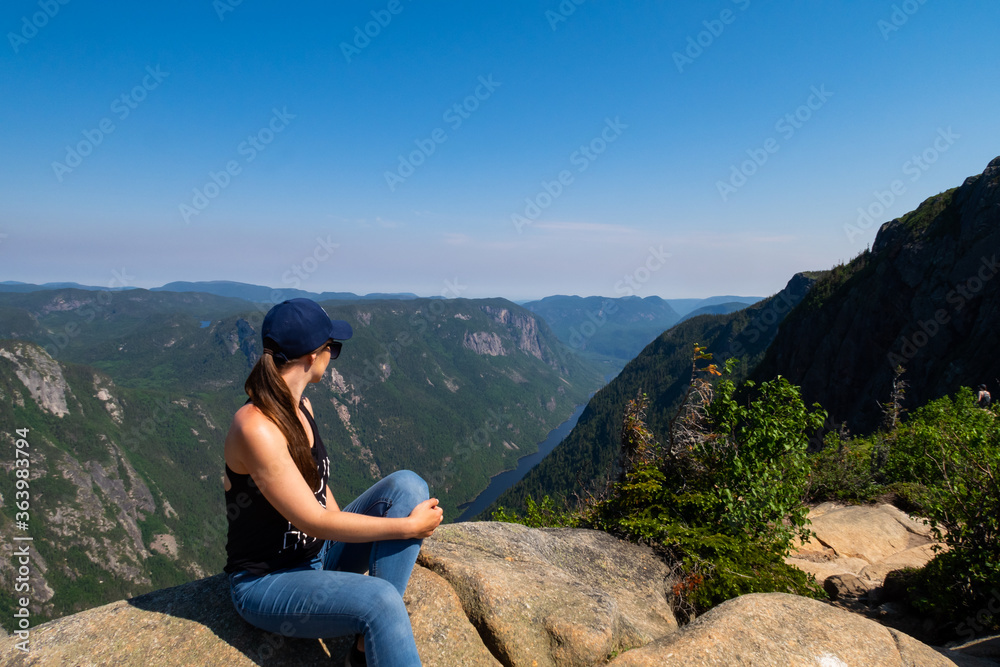 Naklejka premium Young woman standing on the edge of a cliff at the summit of a mountain in the Hautes-gorges-de-la-rivière-Malbaie national park, Canada 