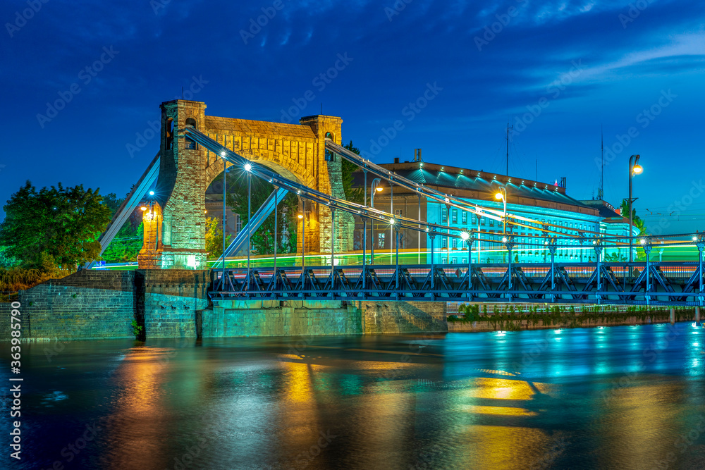 Obraz premium Grunwald Bridge. Grunwaldzki suspension bridge in Wroclaw, Poland at dusk with fast moving cars, trams creating traffic light trails. The most known city landmark seen from the bank side