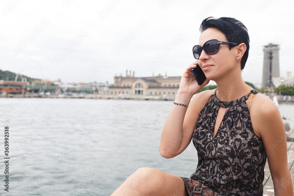 Portrait of a young brunette woman calling with smart phone while enjoying good day outdoors, stylish female tourist talking on her cell telephone while relaxing after walking during summer weekend