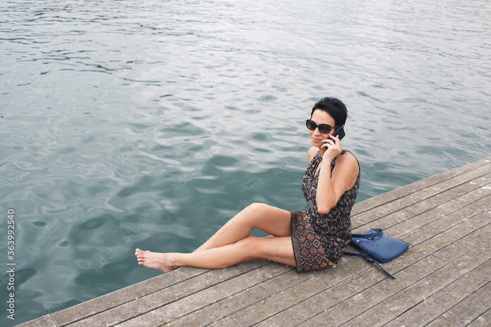 Modern woman having mobile phone conversation while sitting on a pier near sea background with copy space area for your advertising content, female talking on her cellphone while relaxing outside