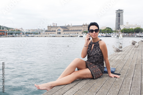 Attractive female calling with smart phone while relaxing after walking during summer vacations, young modern woman having mobile phone conversation while sitting near sea background with copy space