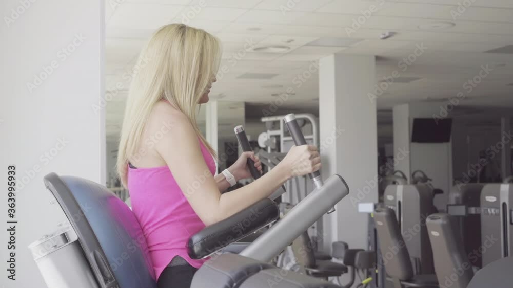 Side view of confident blond woman raising legs in gym. Young beautiful Caucasian sportswoman training muscles on sport inventory. Slim attractive girl exercising on equipment in sports club. Side view of confident blond woman raising legs in gym. Young beautiful Caucasian sportswoman training muscles on sport inventory. Slim attractive girl exercising on equipment in sports club.