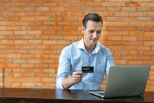 Young business man holding credit card using laptop shopping online in coffee shop cafe with brick wall background