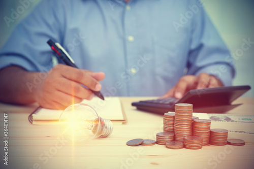 A light bulb and many stacking coins put on the wooden table. Behind blur businessman calculating cash account, expenses, and profits. Concept business, financial, banking, and accounting.