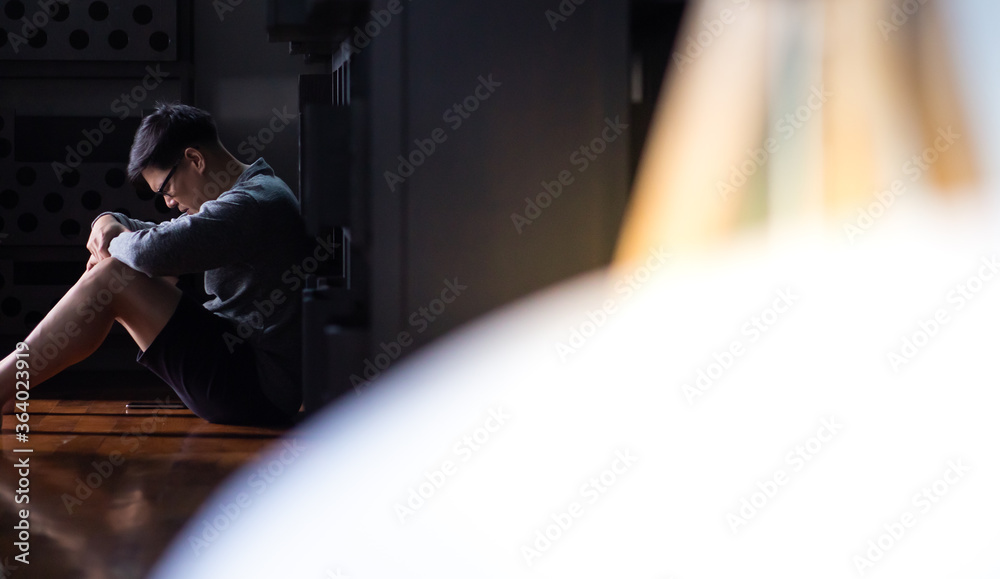 Desperate Asian young man sitting against a wall and looking down at home. Male on black background low key Desperate Asian young man sitting against a wall and looking down at home. Male on black background low key