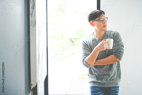 Young Asian man drinking hot coffee while looking through window in living room stay at home. social distancing