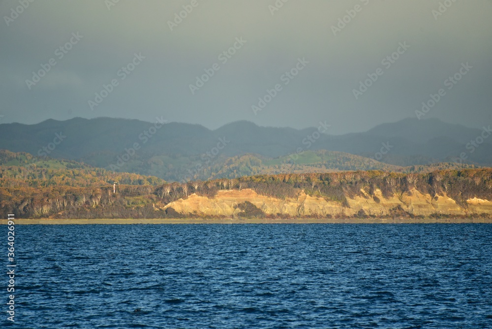 Sakhalin Island coast. View from the sea. Autumn forest on the shore. Clear blue water of the Sea of Okhotsk. Clear autumn day. Northern landscape. 