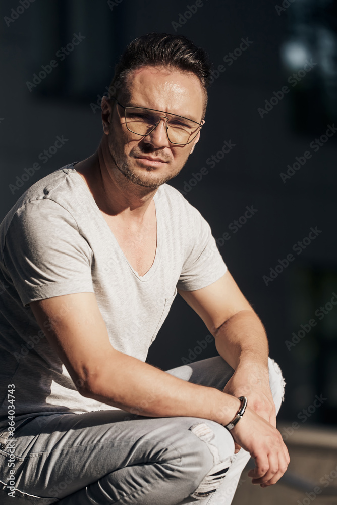 stylish guy with glasses sat on the sidewalk, smiles a little and looks at the camera. strong male gaze. light jeans and a fashionable gray T-shirt. sunlight. close up stylish guy with glasses sat on the sidewalk, smiles a little and looks at the camera. strong male gaze. light jeans and a fashionable gray T-shirt. sunlight. close up