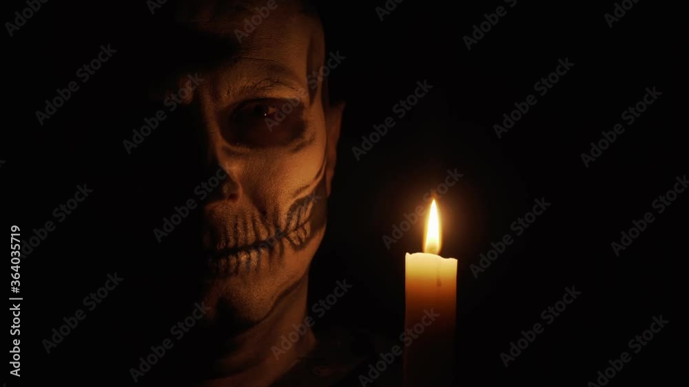 A man with skeleton makeup looks at the camera. A man smiles a creepy and funny smile. Skeleton in the dim light of a candle. Halloween and horror concept. A man with skeleton makeup looks at the camera. A man smiles a creepy and funny smile. Skeleton in the dim light of a candle. Halloween and horror concept.