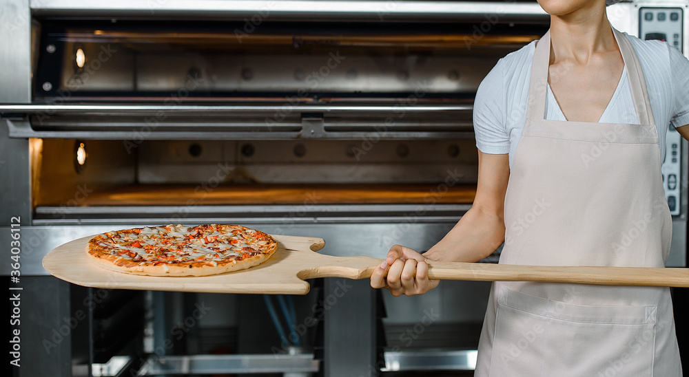 Φωτογραφία Young caucasian woman baker is holding a wood peel with fresh pizza near an oven at a baking manufacture factory