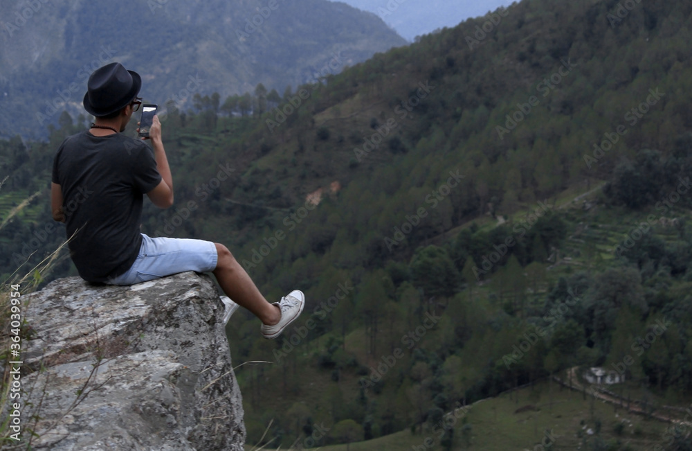 Φωτογραφία Young man wearing a cool hat taking photos on his cellphone of a mountain valley while sitting on the edge of a rock