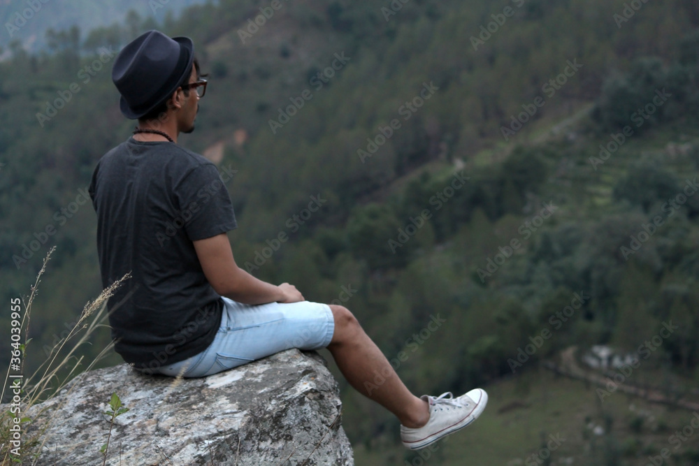 Young man wearing a cool hat sitting on the edge of a rock if front of a beautiful mountain valley.