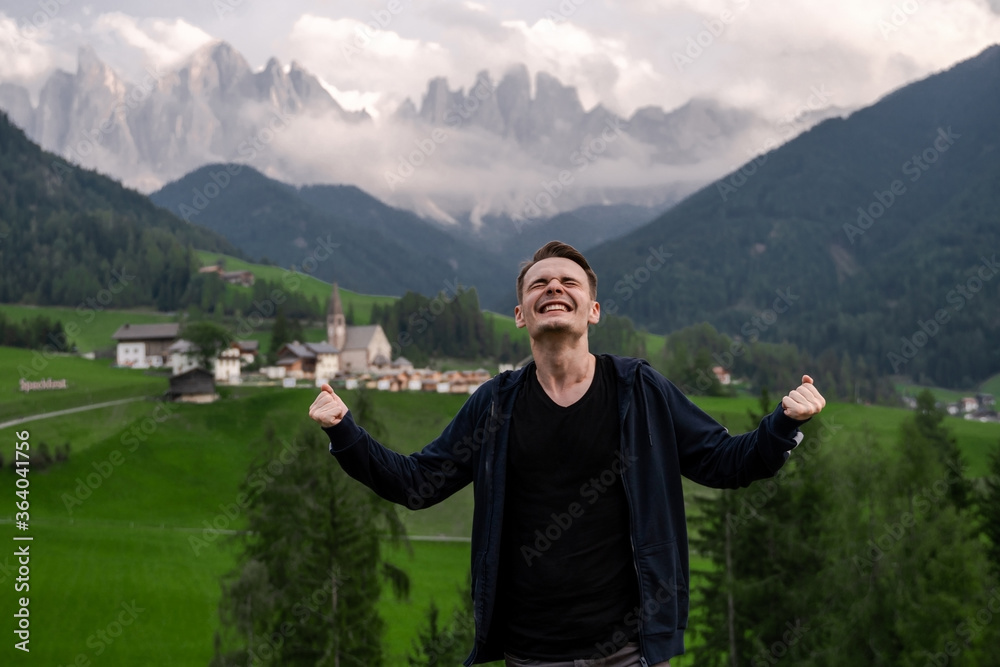 Dolomites. Santa Magdalena. Italy. Smiling, happy brunet man reached the top on background of alpine village, hills, green meadow & high mountains peak wrapped by the clouds in sunny summer day