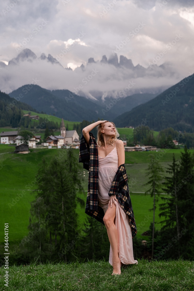 Naklejka premium Dolomites. Santa Magdalena. Italy. Smiling blonde woman in coffee dress & checkered shirt poses on background of alpine village, hills, green meadow & high mountains peak wrapped by the clouds