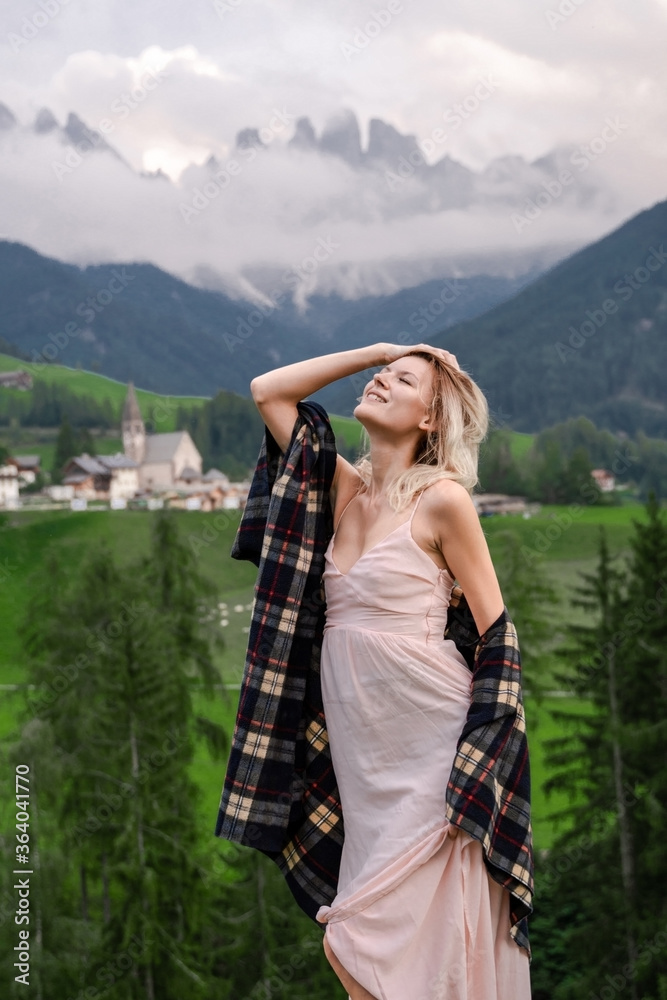 Dolomites. Santa Magdalena. Italy. Blonde woman in coffee dress & checkered shirt raises hand & poses on background of alpine village, hills, green meadow & high mountains peak wrapped by the clouds