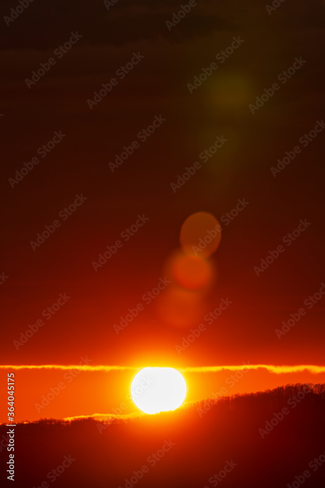 Rising of the sun in mountains, red-orange sun disk rises from tops of mountain range. Beautiful natural lens flare in orange sky, sunbeam light leaks.