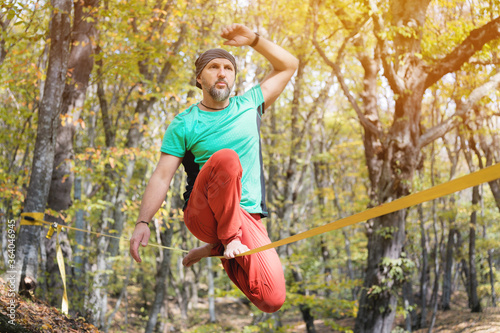 A bearded man aged balancing on a slackline in the autumn forest on a sunny day. The concept of sports leisure at the age of forty