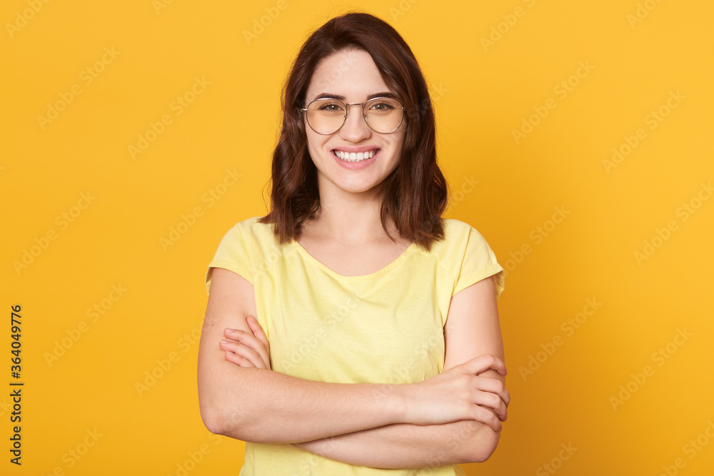 Young attractive brunette girl wearing casual clothing, having beautiful dark hair, looking at camera with toothy smile, lady standing against yellow background with folded hands. Young attractive brunette girl wearing casual clothing, having beautiful dark hair, looking at camera with toothy smile, lady standing against yellow background with folded hands.