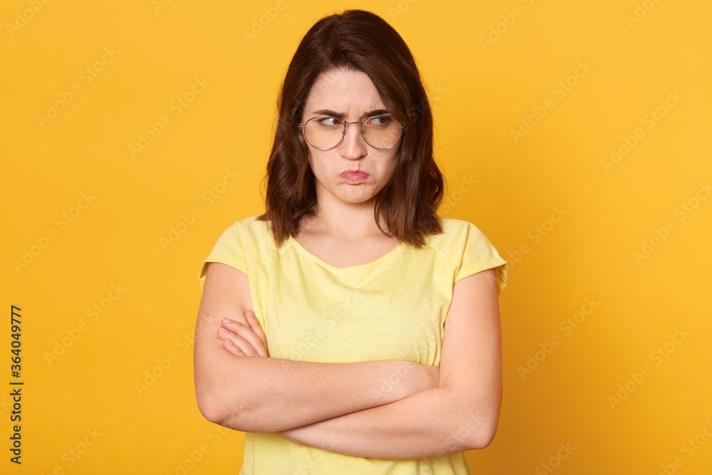 Offended woman wearing yellow t shirt, female with beautiful hair posing with pouty lips, looks upset, standing against yellow wall with folded hands and looking aside. Offended woman wearing yellow t shirt, female with beautiful hair posing with pouty lips, looks upset, standing against yellow wall with folded hands and looking aside.